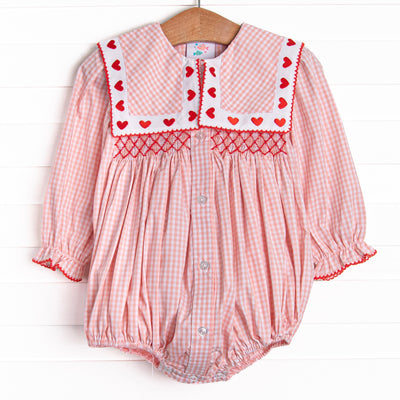  This bubble features pink gingham print, a bib collar with heart embroideries, and a dainty red smocking. 
