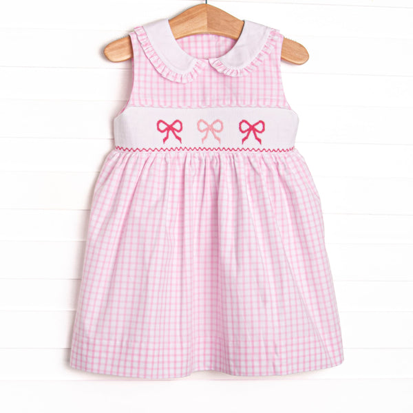 This dress features a pink gingham design, bow smocking, and a ruffle trimmed collar.