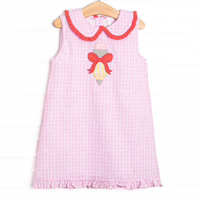  This dress features a pink plaid material, a Peter Pan collar with red ruffle trim, a ruffle bottom edge, and a pencil bow applique. 