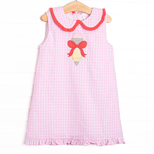  This dress features a pink plaid material, a Peter Pan collar with red ruffle trim, a ruffle bottom edge, and a pencil bow applique. 