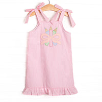 This dress features pink seersucker material, bow straps, a ruffle trimmed edge, and a colored pencils and bow applique.