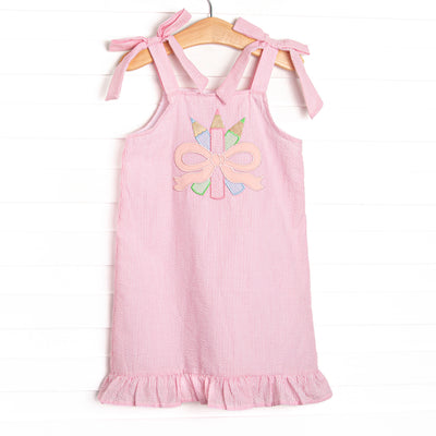 This dress features pink seersucker material, bow straps, a ruffle trimmed edge, and a colored pencils and bow applique.