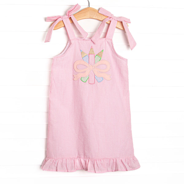 This dress features pink seersucker material, bow straps, a ruffle trimmed edge, and a colored pencils and bow applique.