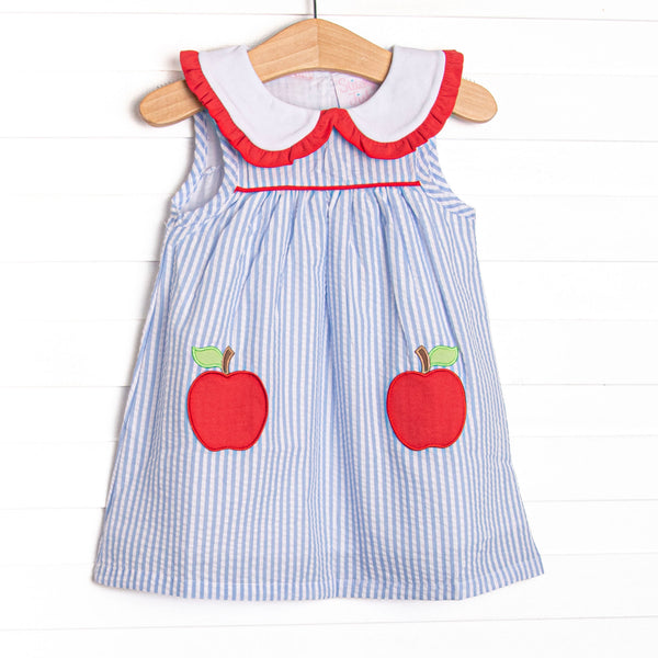 This dress features a blue seersucker print, a peter pan collar with red ruffles, and an apple applique.