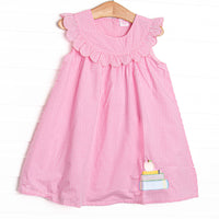  This dress features a pink gingham print, a scalloped bib collar, and an applique of a stack of books topped with an apple. 