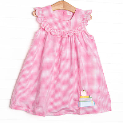  This dress features a pink gingham print, a scalloped bib collar, and an applique of a stack of books topped with an apple. 