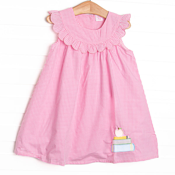  This dress features a pink gingham print, a scalloped bib collar, and an applique of a stack of books topped with an apple. 