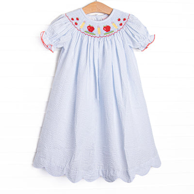 This dress features a blue seersucker print, puff sleeves, and&nbsp; a smocking of apples and pencils.