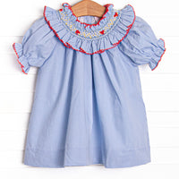 This dress features blue mini gingham fabric, a red ric rac trimmed smocked collar, a bow design, and puff sleeves.