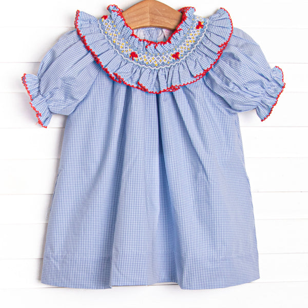 This dress features blue mini gingham fabric, a red ric rac trimmed smocked collar, a bow design, and puff sleeves.