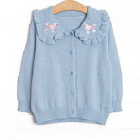  This cardigan features blue knit fabric, pink poodle embroidery, a blue ruffle collar and blue buttons down the front. 