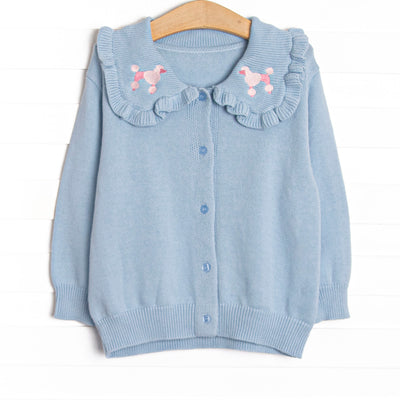  This cardigan features blue knit fabric, pink poodle embroidery, a blue ruffle collar and blue buttons down the front. 