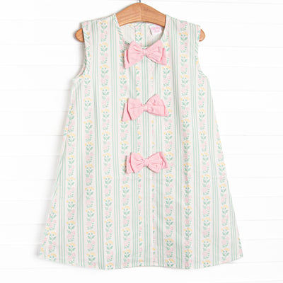 This dress features a green floral print, pink bow details, and button closure on the back.