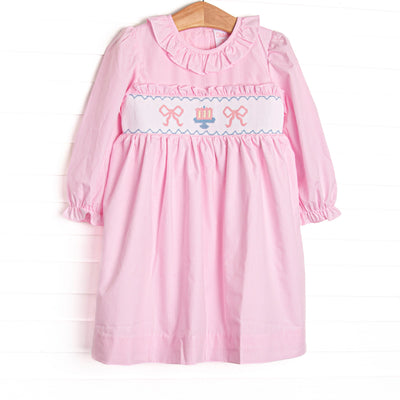 This dress features light pink gingham fabric, bow and cake smocking, ruffle neck trim, and long sleeves.