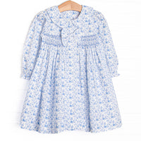 This dress features a blue floral fabric, dainty smocking, a floral peter pan collar, and a pre tied bow.