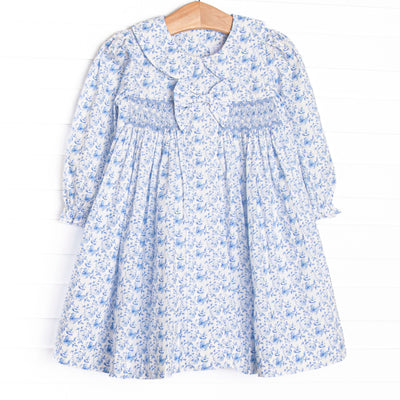 This dress features a blue floral fabric, dainty smocking, a floral peter pan collar, and a pre tied bow.