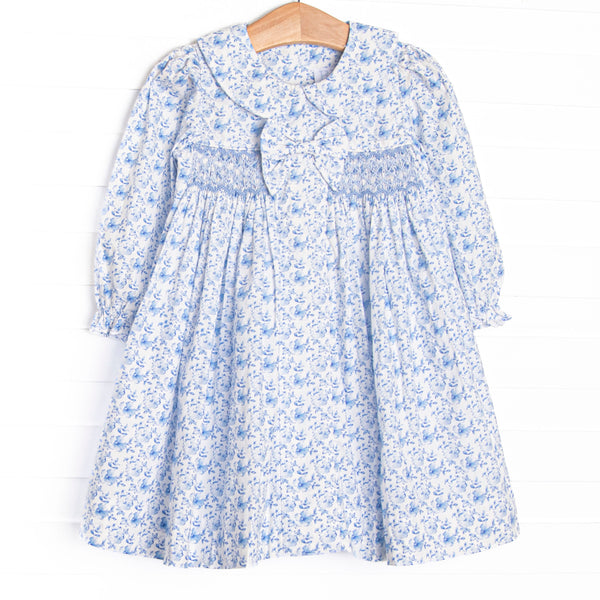 This dress features a blue floral fabric, dainty smocking, a floral peter pan collar, and a pre tied bow.