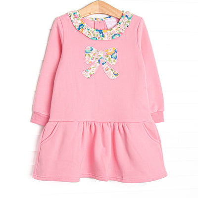 This sweatshirt dress features pink knit fabric, floral ruffle trim, and a floral bow applique. 