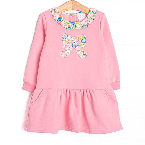 This sweatshirt dress features pink knit fabric, floral ruffle trim, and a floral bow applique. 
