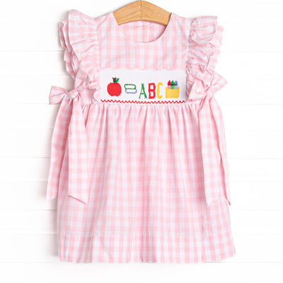  This dress features a pink check print, bows on the sides, and school supplies smocking. 