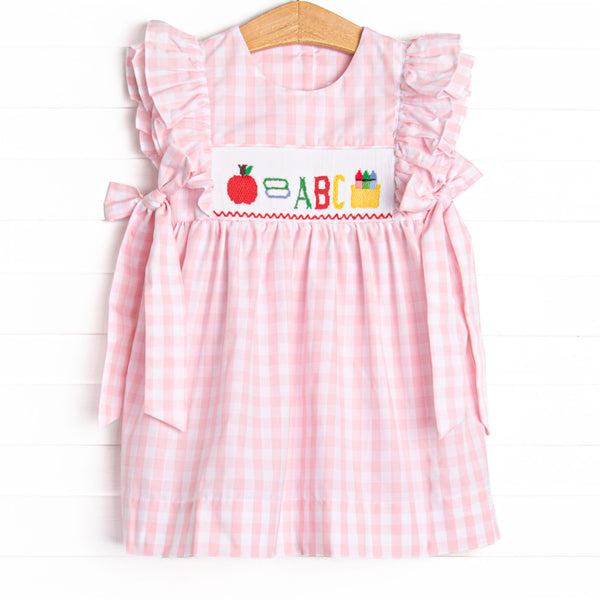  This dress features a pink check print, bows on the sides, and school supplies smocking. 