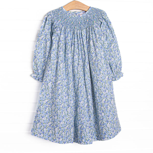 This dress features a blue floral print, a dainty smocking, and buttons down the back.